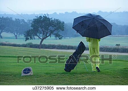 Stock Photography - Rear view of golfer next to golf bag in rain holding umbrella. Fotosearch