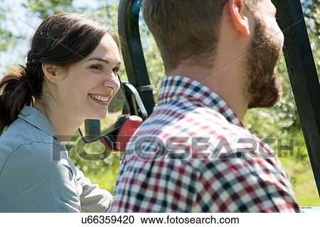 Stock Image - Side view of smiling young couples head and shoulders with tractor. Fotosearch