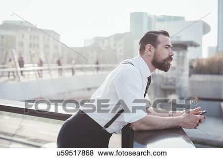 Stock Photo - Stylish businessman with smartphone looking out from office balcony. Fotosearch
