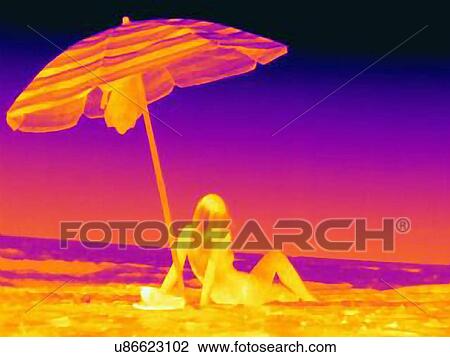 Stock Image - Thermal image rear view of young woman sitting on beach underneath umbrella. Fotosearch