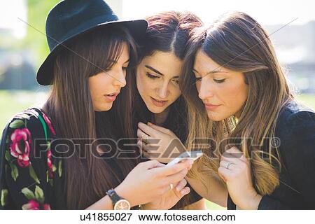 Stock Photo - Three young female friends huddling to share smartphone text message. Fotosearch