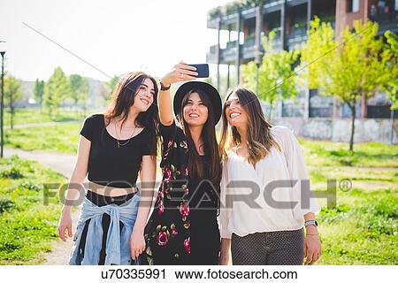 Stock Image - Three young female friends posing for smartphone selfie in park. Fotosearch