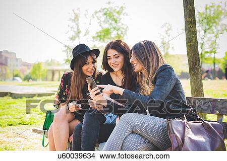 Picture - Three young female friends reading smartphone update on park bench. Fotosearch