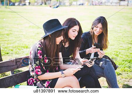 Stock Image - Three young female friends reading their smartphones on park bench. Fotosearch