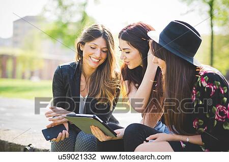 Stock Image - Three young female friends sharing update on digital tablet in park. Fotosearch