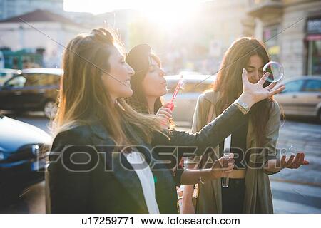 Stock Image - Three young women blowing bubbles on city street. Fotosearch