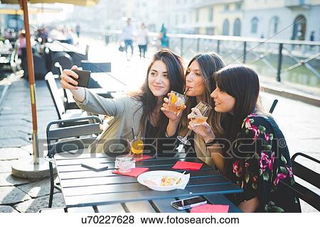 Stock Photo - Three young women posing for smartphone selfie at waterfront cafe. Fotosearch