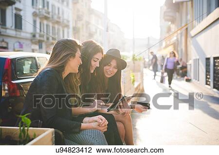 Three young women sitting using digital tablet on city street View Large Photo Image Stock Image - Three young women sitting using digital tablet on city street. Fotosearch
