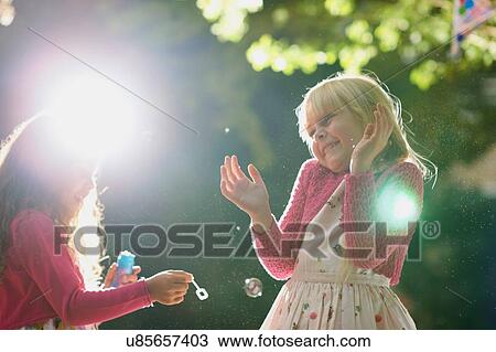 Stock Image - Two girls blowing and popping bubbles in sunlit garden. Fotosearch