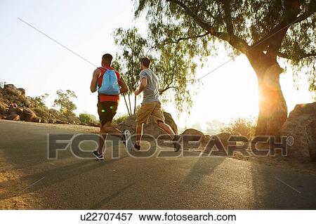 Stock Photo - Two male friends running, outdoors, rear view. Fotosearch