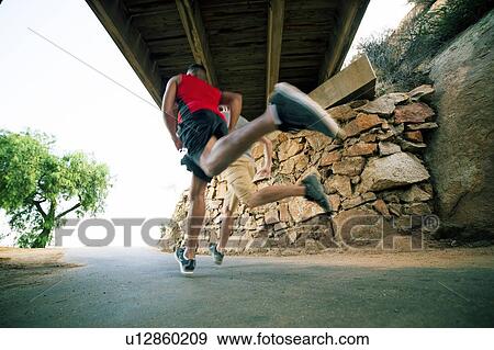 Stock Photo - Two male friends, running under bridge, rear view, low angle view. Fotosearch