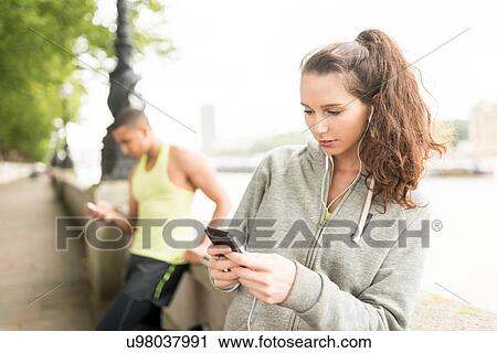 Stock Image - Two runners texting on smartphone at riverside. Fotosearch