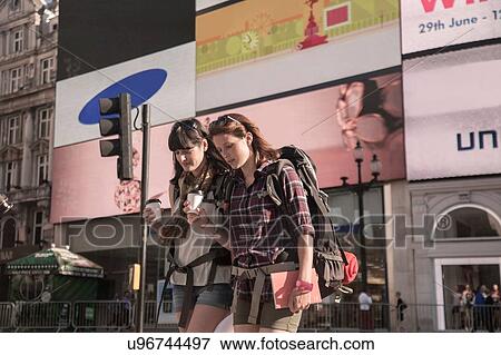 Stock Photo - Two women backpackers strolling with takeaway coffee, Picadilly Circus, London, UK. Fotosearch