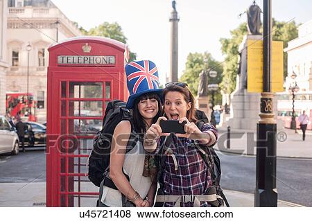 Stock Photo - Two women backpackers taking smartphone selfie at red telephone box, London, UK. Fotosearch