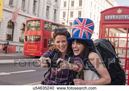 Stock Image - Two women backpackers wearing union jack hat taking smartphone selfie, London, UK. Fotosearch