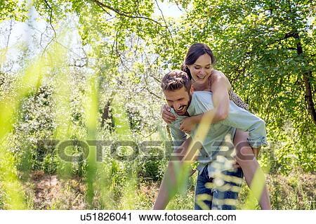 Stock Image - View through foliage of young man giving young woman piggyback. Fotosearch