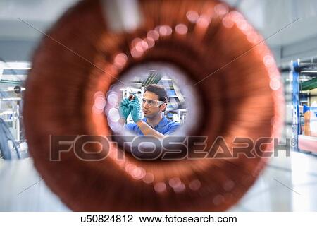 Stock Image - Worker inspecting electromagnetic coil seen through large coil in electromagnetics factory. Fotosearch