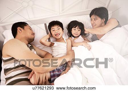 Stock Image - Young Chinese family of parents and two young children laying in bed together at home. Fotosearch