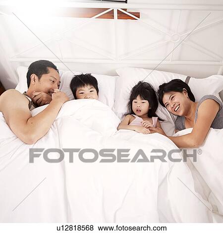 Stock Photo - Young Chinese family of parents and two young children laying in bed together at home. Fotosearch