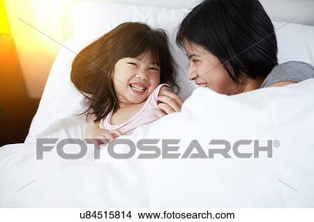 Picture - Young Chinese mother and daughter laying in bed together at home. Fotosearch