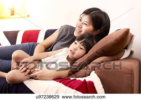 Stock Photo - Young Chinese mother and daughter laying on sofa watching the television together at home. Fotosearch