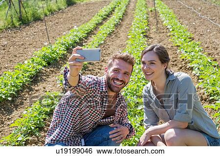 Stock Photograph - Young couple crouched in vegetable garden using smartphone to take selfie. Fotosearch