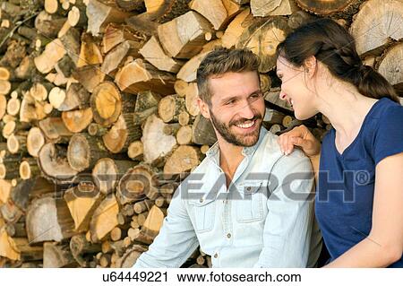 Stock Image - Young couple sitting in front of chopped wood face to face smiling. Fotosearch