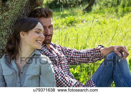 Stock Photo - Young couple sitting leaning against tree together. Fotosearch