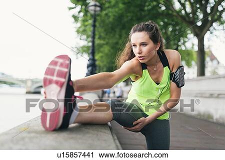 Stock Image - Young female runner touching toes on riverside. Fotosearch