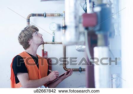 Stock Image - Young man in boiler room holding clipboard inspecting heating system, looking up. Fotosearch