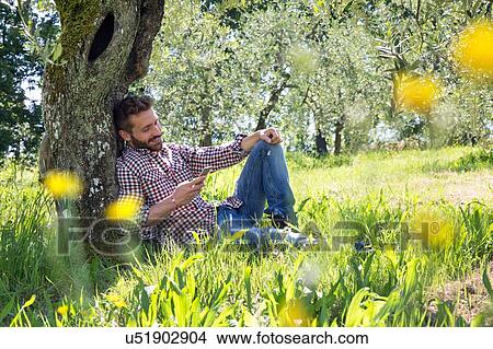 Young man sitting leaning against tree using smartphone looking down smiling View Large Photo Image Picture - Young man sitting leaning against tree using smartphone looking down smiling. Fotosearch