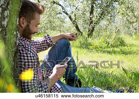 Young man sitting leaning against tree using smartphone looking down smiling View Large Photo Image Stock Photography - Young man sitting leaning against tree using smartphone looking down smiling. Fotosearch