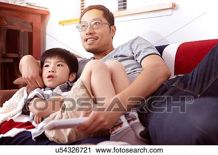 Stock Image - Young, modern Chinese family of father and young son sitting on sofa watching television together at home. Fotosearch