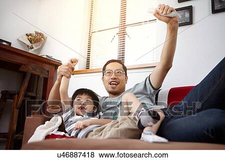 Stock Photo - Young, modern Chinese family of father and young son sitting on sofa watching and cheering at television together at home. Fotosearch