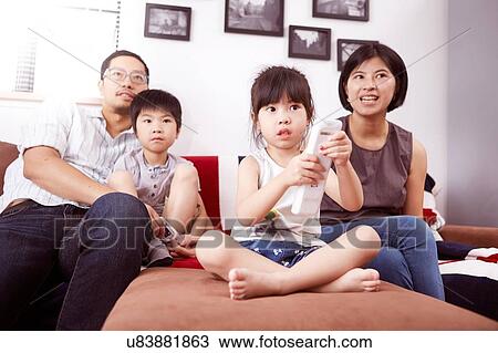 Stock Image - Young, modern Chinese family of parents and two young children sitting on sofa watching television together at home. Fotosearch