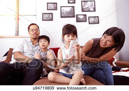 Young, modern Chinese family of parents and two young children sitting on sofa watching television together at home View Large Photo Image Stock Photo - Young, modern Chinese family of parents and two young children sitting on sofa watching television together at home. Fotosearch