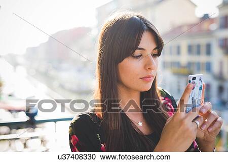 Stock Image - Young woman reading smartphone texts on waterfront balcony. Fotosearch