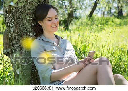 Young woman sitting leaning against tree using smartphone looking down smiling View Large Photo Image Stock Image - Young woman sitting leaning against tree using smartphone looking down smiling. Fotosearch