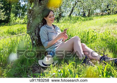 Young woman sitting leaning against tree using smartphone looking down smiling View Large Photo Image Stock Photo - Young woman sitting leaning against tree using smartphone looking down smiling. Fotosearch