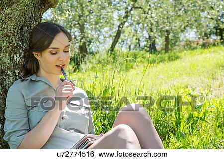 Young woman sitting leaning against tree, looking down chewing on pen View Large Photo Image Stock Photograph - Young woman sitting leaning against tree, looking down chewing on pen. Fotosearch