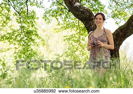 Young woman standing leaning against tree holding camera looking away smiling View Large Photo Image Stock Image - Young woman standing leaning against tree holding camera looking away smiling. Fotosearch