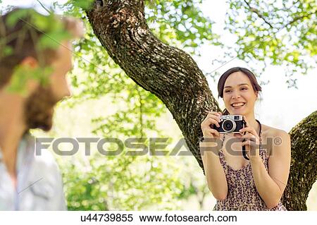 Stock Photography - Young woman underneath tree taking photograph of young man. Fotosearch
