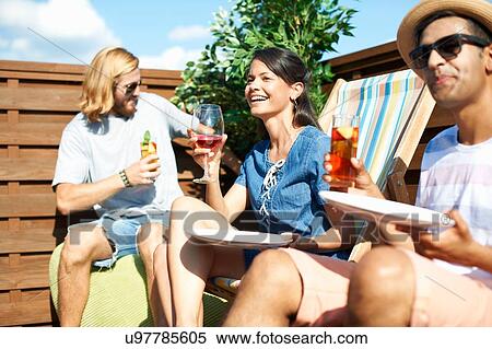 Stock Photography - Adult friends sitting on deck chairs drinking at rooftop party. Fotosearch