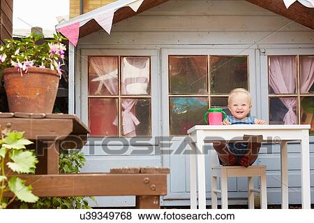 Stock Photo - Baby girl sitting at table in front of playhouse looking at camera smiling. Fotosearch