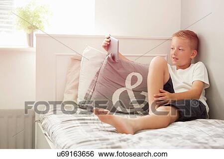 Boy sitting on bed holding digital tablet looking away View Large Photo Image Stock Photograph - Boy sitting on bed holding digital tablet looking away. Fotosearch