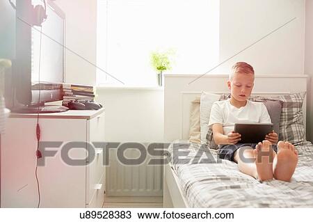Stock Image - Boy sitting on bed looking down at digital tablet. Fotosearch