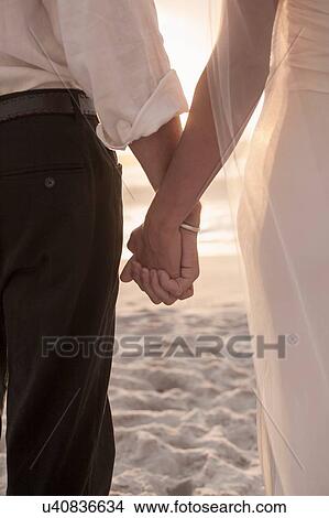 Picture - Bride and groom holding hands on beach against sunset. Fotosearch