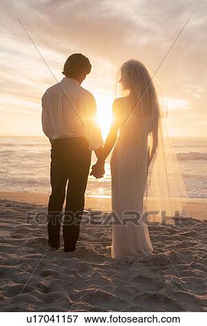 Stock Photo - Bride and groom holding hands on beach against sunset. Fotosearch