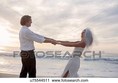 Stock Image - Bride and groom on beach, holding hands, fooling around, laughing. Fotosearch