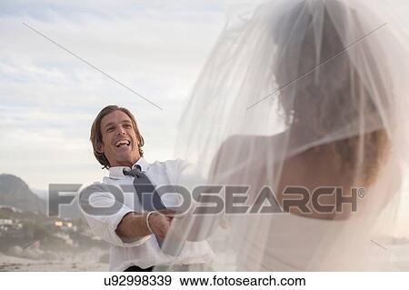 Stock Photo - Bride and groom on beach, holding hands, fooling around, laughing. Fotosearch